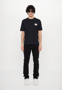 Tricou din bumbac negru cu mâneci scurte și un mic patch cu logo argintiu pe piept, purtat cu blugi negri și adidași albi.
