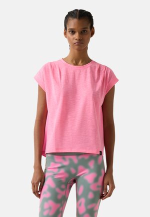 Frau mit geflochtenem Haar, die ein lockeres rosa Kurzarmshirt und graue Leggings mit pinken abstrakten Mustern trägt, vor einem weißen Hintergrund stehend.