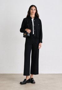 Chaqueta negra corta con cierres de botón, combinada con pantalones de tiro alto de pierna ancha negros y mocasines negros; la modelo sostiene un bolso pequeño.