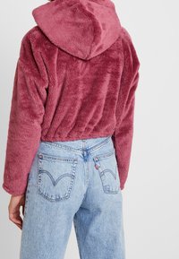 Mujer vistiendo una chaqueta corta de felpa burdeos con capucha combinada con jeans de mezclilla azul claro de cintura alta, vista desde atrás.