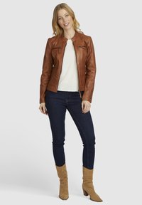 Bruine leren jas met ritszakken, gestructureerd ontwerp, ronde kraag, gecombineerd met een crèmekleurige top en donkere jeans, met beige enkellaarsjes.