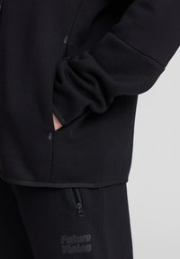 Felpa nera con una texture liscia, dotata di una tasca con zip e un logo. Ha una vestibilità rilassata e bordature a contrasto lungo i lati.