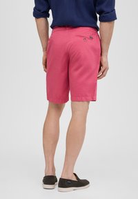 Shorts roses pour hommes avec poche arrière et détail de bouton. Fabriqués en tissu léger avec une texture lisse. Portés avec des chaussures noires à enfiler.