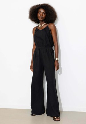 Femme avec une coiffure afro naturelle portant une combinaison noire sans manches, un collier en coquillage, un bracelet en or et des sandales à imprimé léopard, debout contre un mur blanc.