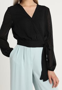 Blouse portefeuille noire en tissu texturé, manches longues et taille nouée. Associée à un pantalon large bleu clair.