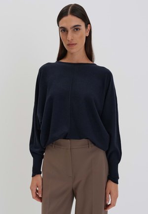 Maglione oversized blu navy con scollo a barca ampio e spalle scese, realizzato in tessuto morbido. Abbinato a pantaloni tailleur beige chiaro.