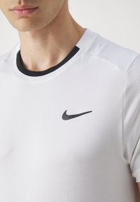 Hvit atletisk skjorte med svart krage og en liten svart Nike-logo på brystet, laget av et teksturert, pustende stoff.