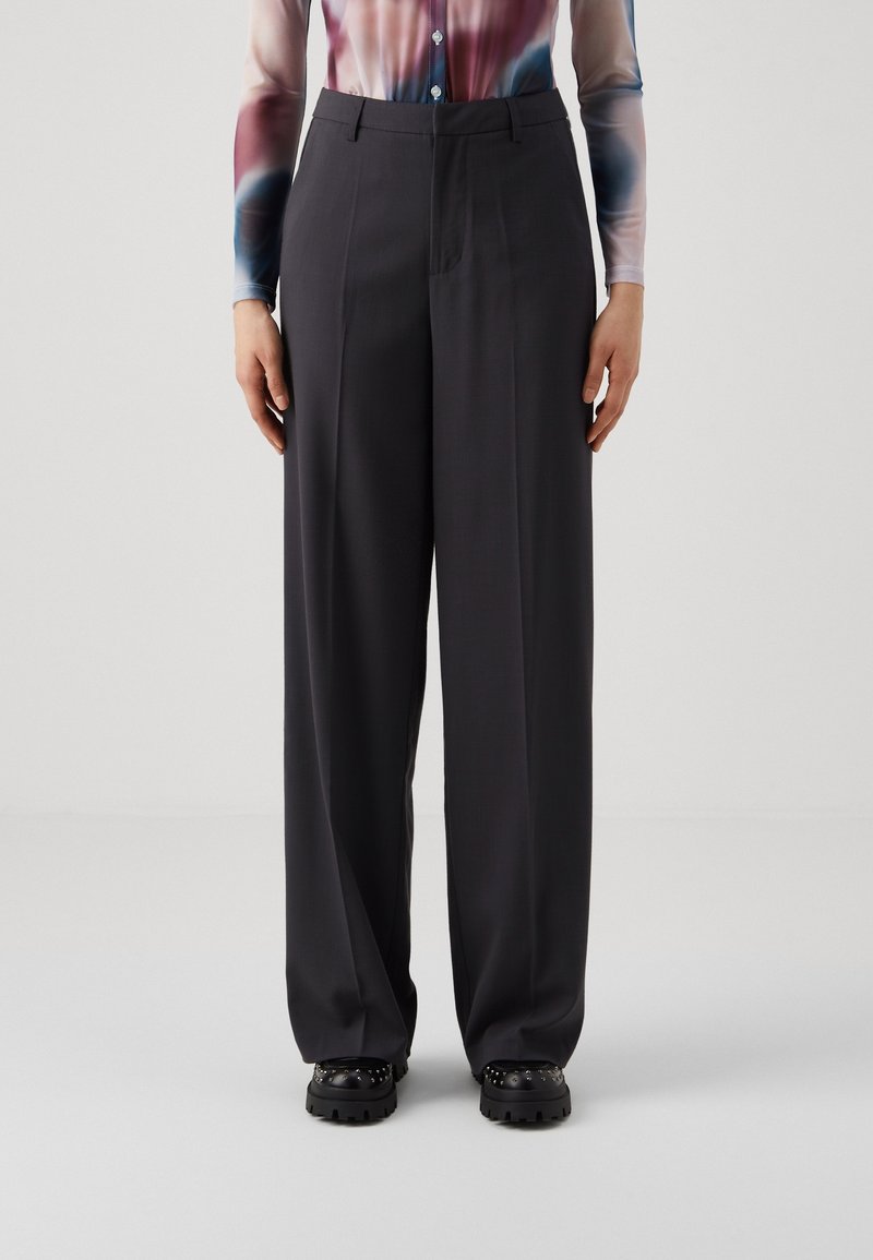 Personne portant un pantalon noir taille haute à jambes larges, un t-shirt manches longues tie-dye et des chaussures noires cloutées, debout devant un fond uni.