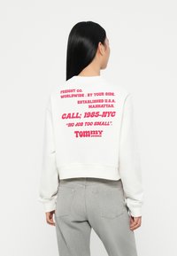 Witte cropped sweatshirt met rode grafische tekst op de achterkant. De tekst omvat de merknaam en de vestigingslocatie. De stof is soepel en heeft een casual pasvorm.