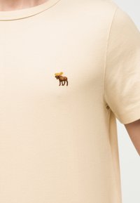 Μπεζ κοντομάνικη μπλούζα με μικρό κεντημένο καφέ αλκάνι στην αριστερή πλευρά του στήθους.