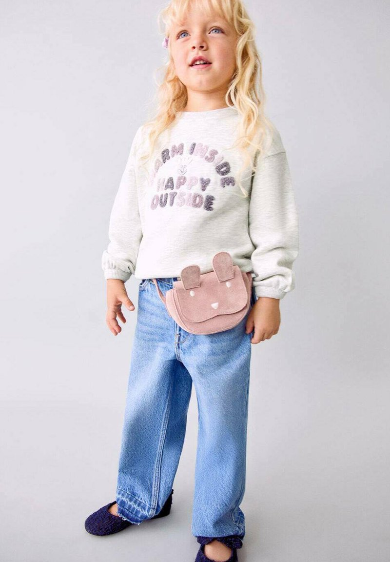Bambino con lunghi capelli biondi che indossa una felpa chiara con del testo, jeans blu, pantofole scure e una borsa a vita a forma di animale rosa.