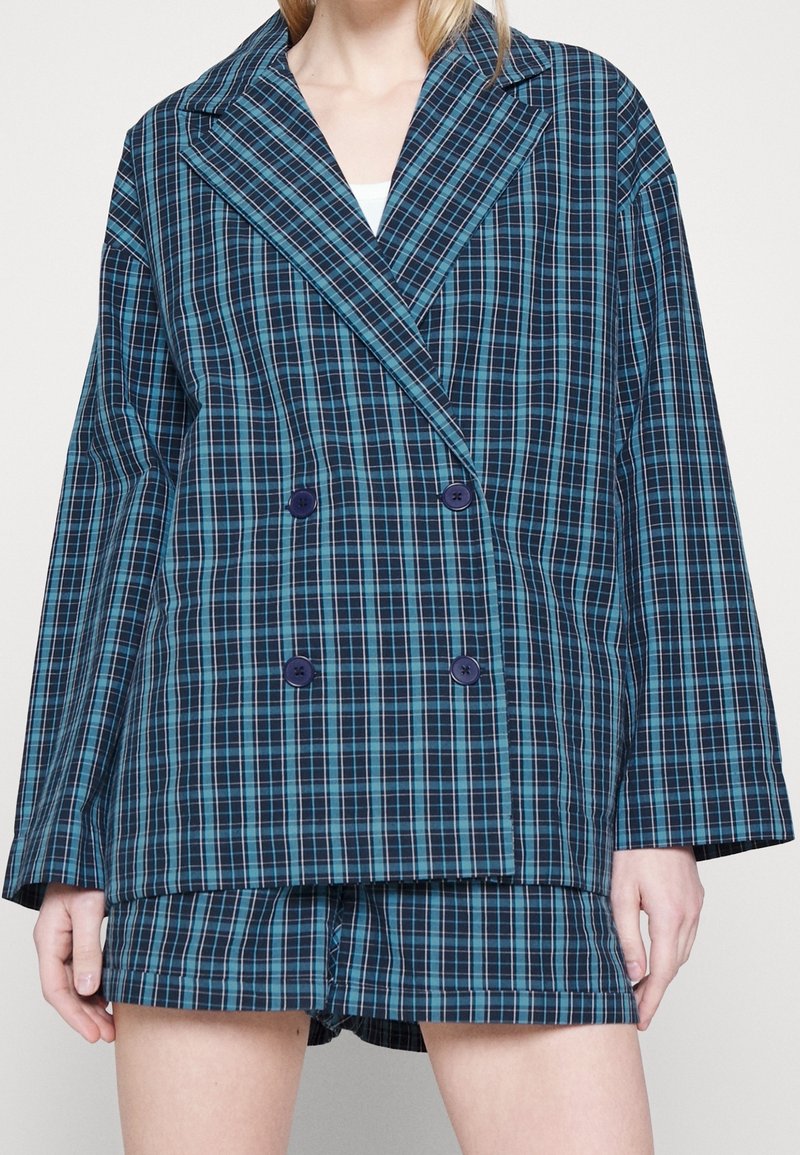 Kobieta ubrana w niebiesko-czarną kratowaną marynarkę zapinaną na dwa rzędy guzików oraz dopasowane szorty, stojąca na gładkim tle.