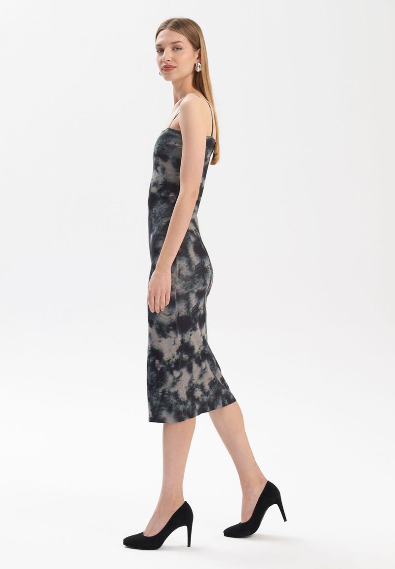 Dopasowana, bezrękawnikowa sukienka midi w ciemnym wzorze tie-dye, wykonana z gładkiego materiału. Ma boczne rozcięcie i zestawiona z czarnymi szpilkami.