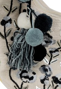 Tessuto ricamato con motivi floreali; presenta pompon e frange blu, neri e bianchi; sfondo beige con accenti metallici.