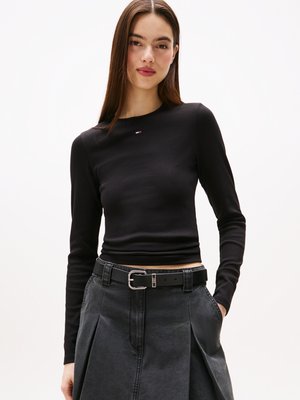 Top preto de manga longa com um tecido texturizado, apresentando um pequeno detalhe de logotipo. Combinado com uma saia cinza e um cinto preto com fivela de metal.