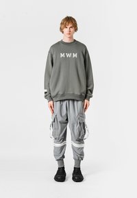 Sudadera gris con letras "MWM", combinada con pantalones cargo grises que tienen bolsillos laterales y acentos en blanco contrastantes. Footwear negro completa el look.