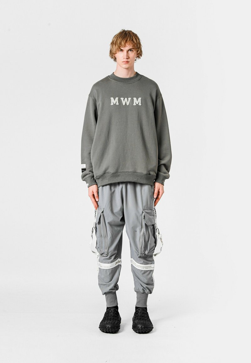 Sudadera gris con letras "MWM", combinada con pantalones cargo grises que tienen bolsillos laterales y acentos en blanco contrastantes. Footwear negro completa el look.