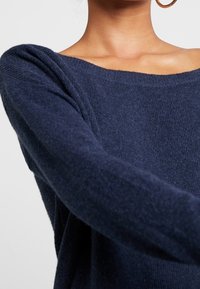 Pull en maille bleu marine avec un large décolleté et des manches longues. Tissu texturé, coupe décontractée, et détail d'épaule légèrement tombante.