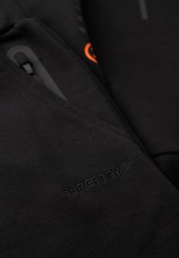 Zwarte hoodie met rits, voorzien van geborduurd logo in bijpassend garen, textuurstof en oranje accent op de rits.
