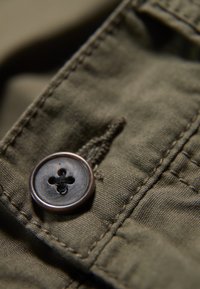 Botón marrón con un diseño de cuatro agujeros sobre una tela verde oliva, que presenta costuras visibles y una superficie texturizada.