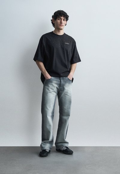 Νεαρός με σγουρά μαλλιά στέκεται με τα χέρια στις τσέπες, φορώντας μαύρο oversize T-shirt με λευκές βούλες, ξεβαμμένο ανοιχτό μπλε τζιν και μαύρα αθλητικά παπούτσια.
