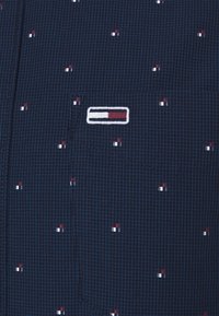 Tissu bleu foncé avec de fines lignes de quadrillage bleu clair, petits motifs carrés rouges et blancs, et un logo rectangulaire cousu en rouge, blanc et bleu.