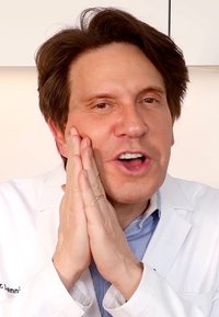 En man som bär en vit labbrock och en blårandig skjorta håller sina händer tillsammans nära ansiktet och uttrycker entusiasm eller engagemang.