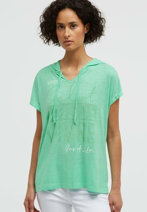 Frau mit lockigem Haar, die ein hellgrünes Kurzarm-Hoodie mit französischem Text und weiße Hose trägt, steht vor einem einfarbigen Hintergrund.