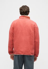 Άνδρας που φορά ροδακινί φλις ζακέτα και σκούρο γκρι παντελόνι, απεικονίζεται από πίσω μπροστά σε απλό φωτεινό φόντο.