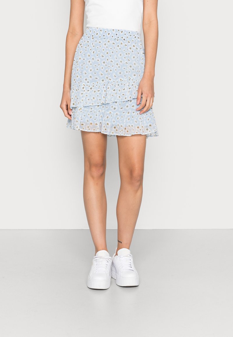 Mini-jupe à volants bleu clair à motif floral portée avec des baskets blanches et un haut blanc, montrée de la mi-cuisse aux pieds sur un fond uni.