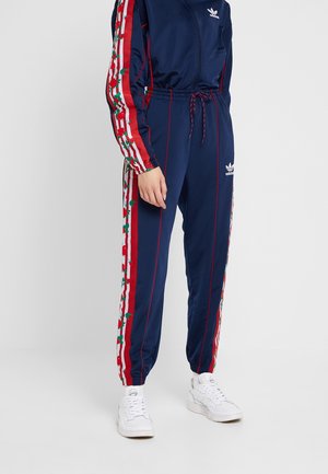 Personne portant un pantalon et une veste de survêtement Adidas bleu marine avec des bandes latérales florales rouges et blanches et des baskets blanches sur un fond uni.