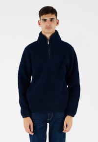 Pullover in pile blu navy con colletto alto e chiusura a mezza zip. Presenta un tessuto testurizzato e polsini elasticizzati. Indossato con jeans scuri.