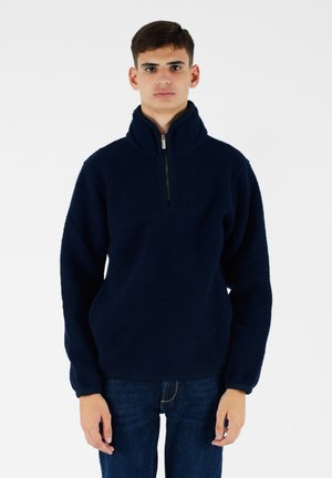 Pullover in pile blu navy con colletto alto e chiusura a mezza zip. Presenta un tessuto testurizzato e polsini elasticizzati. Indossato con jeans scuri.