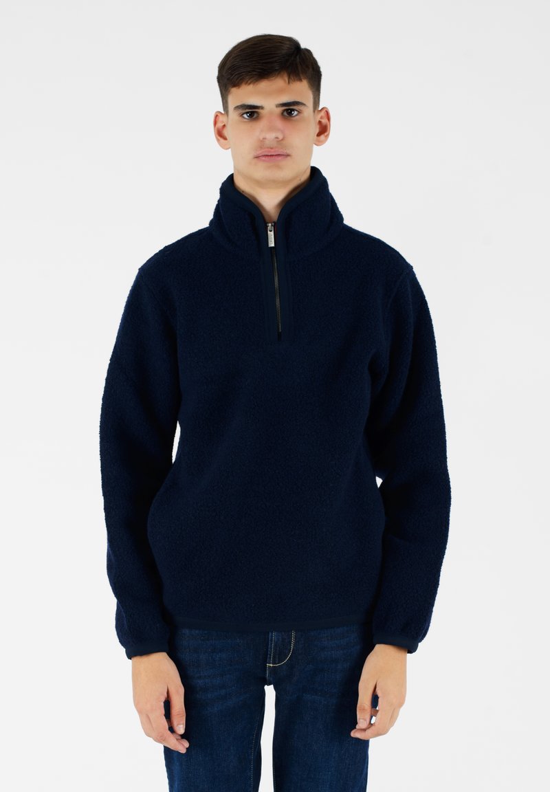Pullover in pile blu navy con colletto alto e chiusura a mezza zip. Presenta un tessuto testurizzato e polsini elasticizzati. Indossato con jeans scuri.