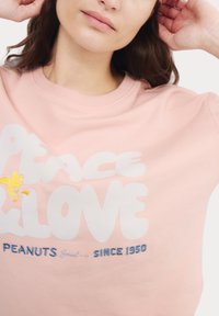 Felpa rosa con grande testo bianco "PEACE LOVE" e grafica gialla di Snoopy, accompagnata dal dettaglio blu "PEANUTS Social Club SINCE 1950".