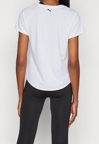 Camiseta atlética blanca con mangas cortas, cuello redondo y tejido texturizado. Presenta un logo negro en la parte posterior, combinada con leggings negros.