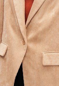 Veste en velours côtelé beige avec une surface texturée, un revers cranté, une fermeture à un bouton et deux poches avant. La doublure révèle un tissu rouge en dessous.