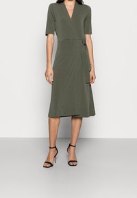 Olivgrön omslagsklänning med korta ärmar, knälång och med ett självknutet bälte i midjan, parat med svarta remsandaler med hög klack.