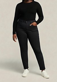 Person wearing black long-sleeve top, fitted black pants with hand in pocket, and white sneakers, standing against a plain background.