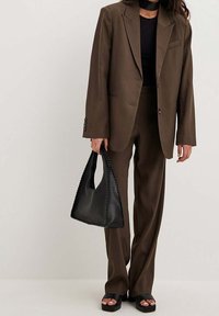 Brązowy garnitur szyty na miarę o luźnym kroju, w zestawie z czarną torbą z plecionym brzegiem. Czarny top i masywne czarne sandały dopełniają całość.