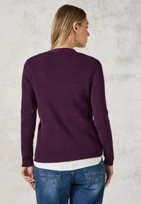 Rippenshirt in Lila mit langen Ärmeln und Rundhalsausschnitt, kombiniert mit einem weißen Hemd am Saum, getragen mit blauen Jeans.