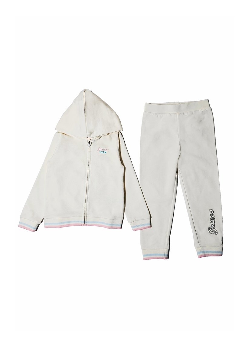 Tuta in pile bianco composto da una felpa con zip e maniche con strisce arcobaleno e pantaloni abbinati. I pantaloni hanno la scritta "Guess" ricamata sulla gamba.