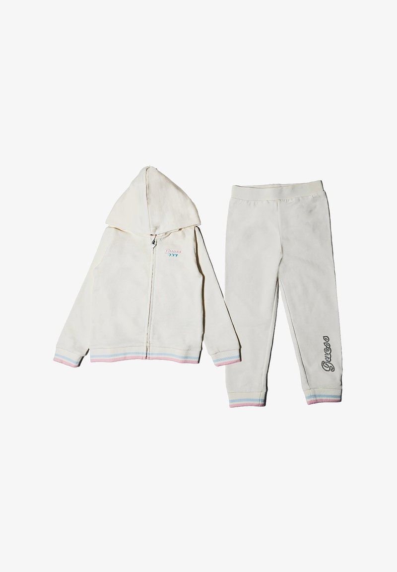 Tuta in pile bianco composto da una felpa con zip e maniche con strisce arcobaleno e pantaloni abbinati. I pantaloni hanno la scritta "Guess" ricamata sulla gamba.