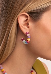 Gros plan d'une oreille portant une boucle d'oreille en forme d'anneau ornée de perles colorées roses, violettes, jaunes, bleues et vertes, sur fond de cheveux blonds.