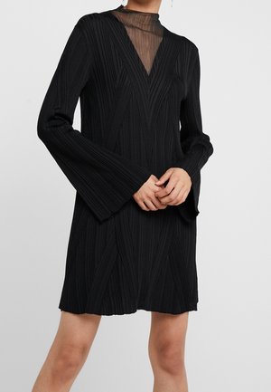 Kobieta ubrana w czarną, prążkowaną, długą sukienkę z rozszerzanymi mankietami i przezroczystą wstawką z wysokim kołnierzem, stoi z złączonymi rękami.