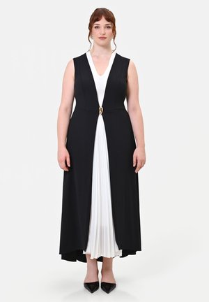 Donna che indossa un cappotto nero senza maniche con chiusura centrale dorata sopra un vestito bianco plissettato e scarpe nere appuntite, in piedi su uno sfondo bianco.