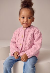 Cardigan rosa lavorato a maglia con un motivo a trecce testurizzato, caratterizzato da bottoni in legno e collo tondo, abbinato a jeans di denim blu.