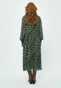 Langes, fließendes Kleid mit einem grünen und blauen abstrakten Muster, transparenter Stoff und elastische Ärmel; kombiniert mit knielangen olivgrünen Stiefeln.