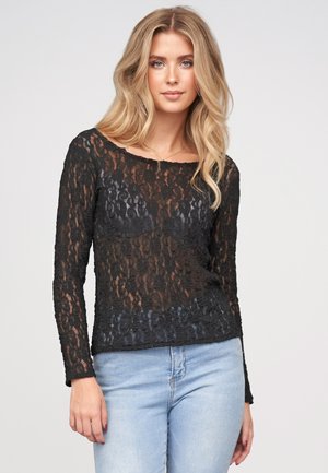 Jeune femme aux longs cheveux blonds portant un haut noir en dentelle transparente à manches longues et un jean bleu clair sur un fond uni.