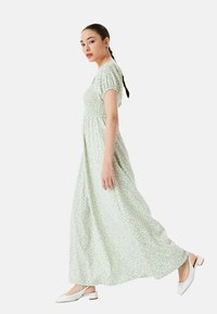Grøn blomstret maxi-kjole med korte puffærmer og smocket liv. Båret med hvide slip-on sko, poserer modellen i profil på en ensfarvet baggrund.